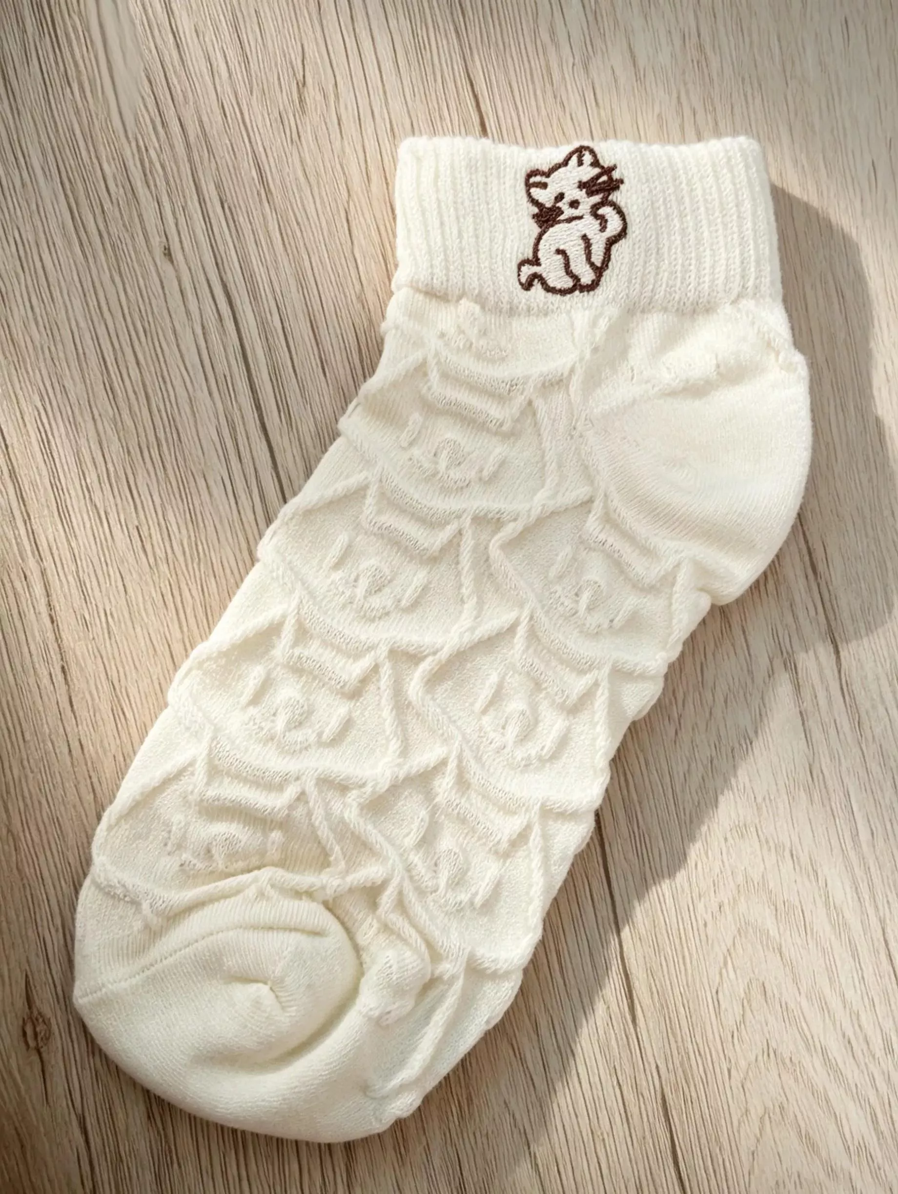 單隻白色針織襪平放於木質桌面上，展示精緻的立體織紋與襪口小貓刺繡。