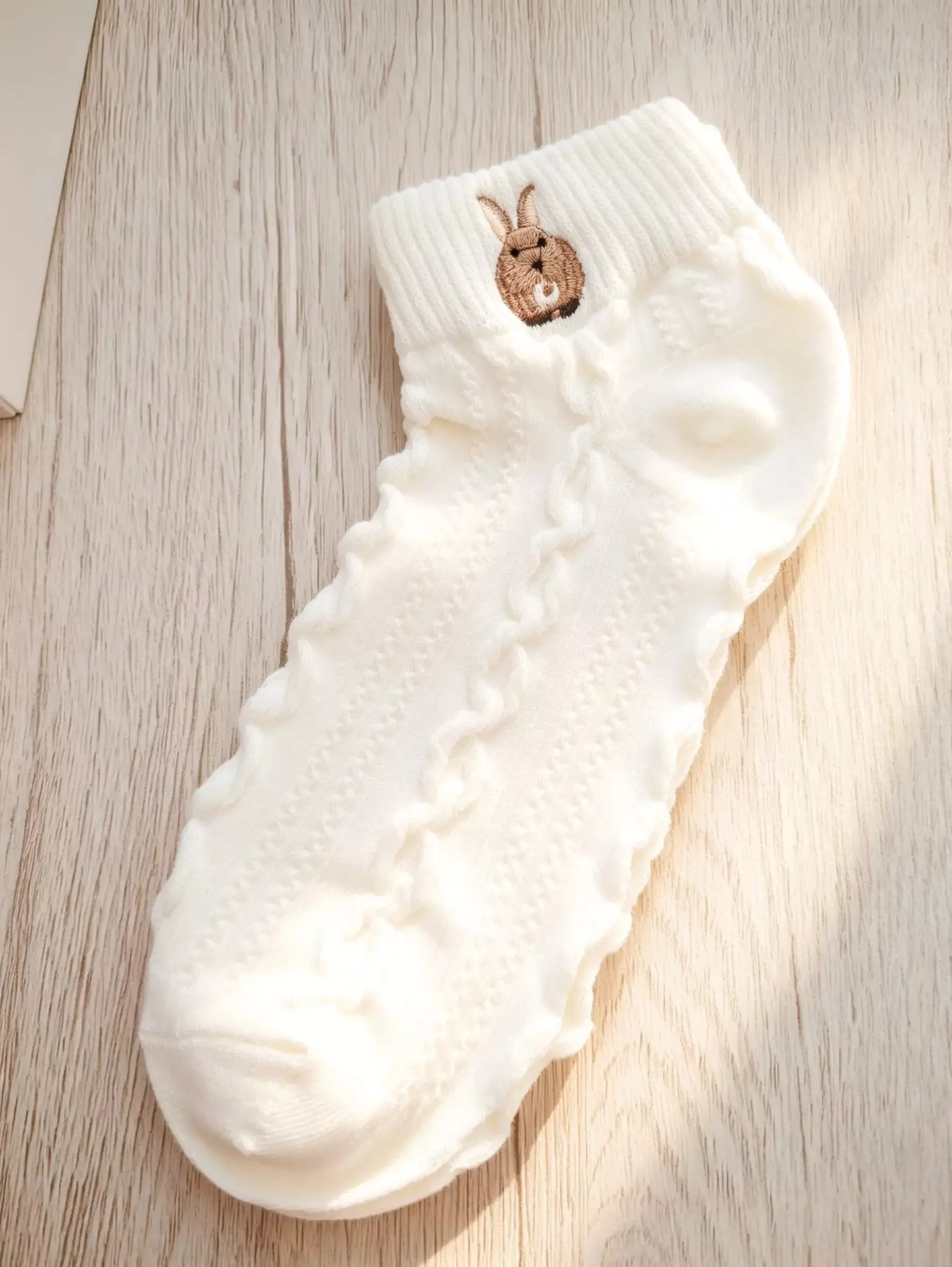 白色針織中筒襪，襪口處有棕色小兔子刺繡，細緻紋路設計。